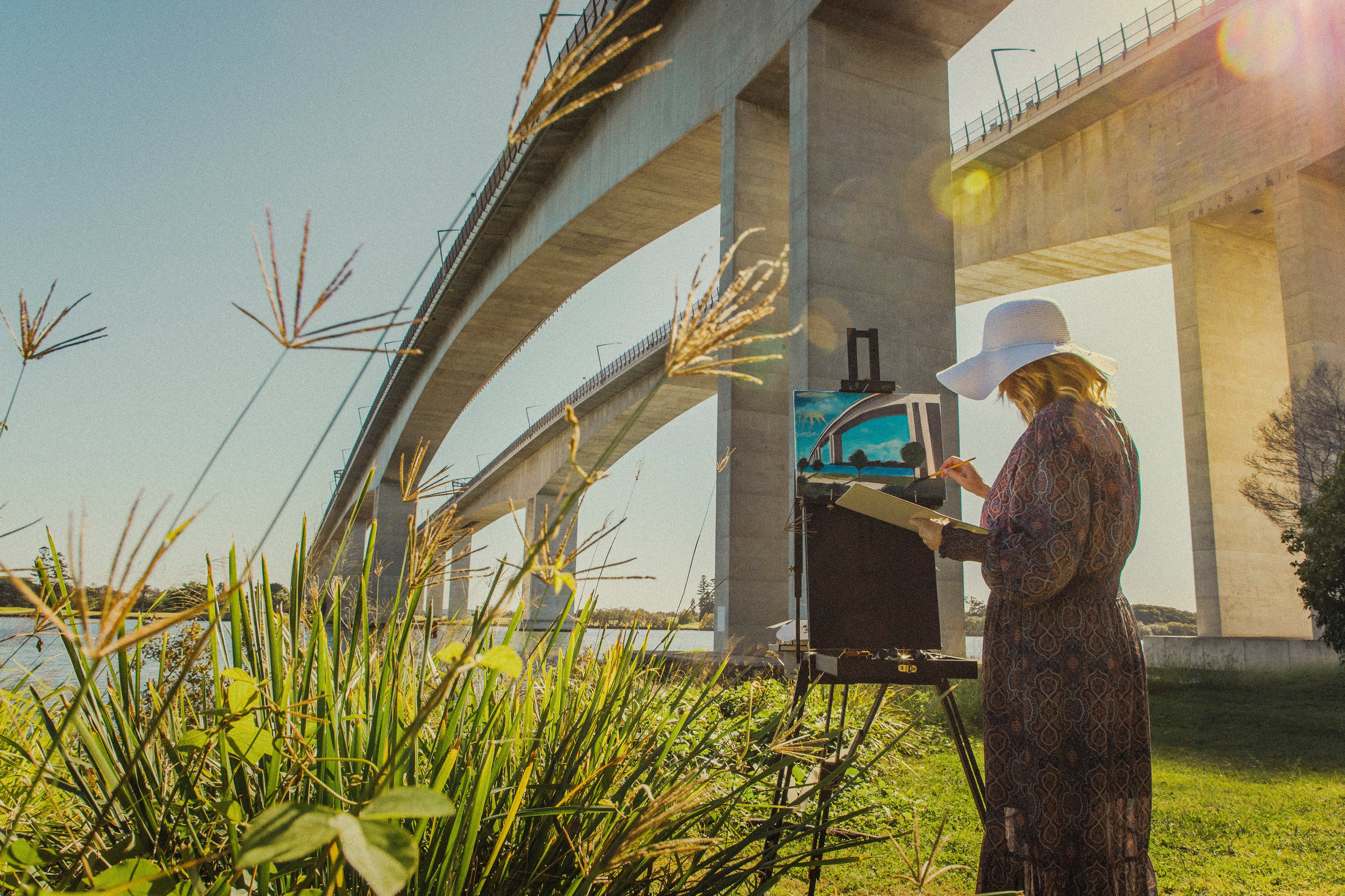 Person in a hat painting by the water under a bridge